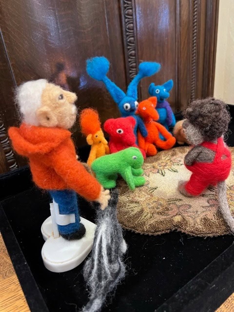 a felted wool figure in an orange hoodie faces a group of colorful felt creatures, while a smaller figure in red stands nearby on a patterned cushion.