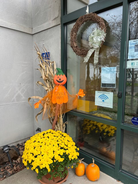 a scarecrow with "pumpkin happy" sign, yellow chrysanthemums, two pumpkins, and corn stalks are arranged by a glass door decorated with a grapevine wreath.