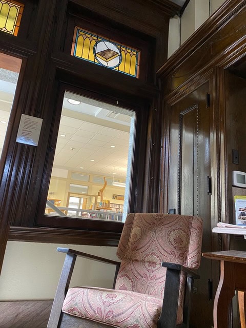 an upholstered armchair sits next to a small wooden table in a room with dark wood trim, stained glass, and a window looking into a brightly lit interior space.