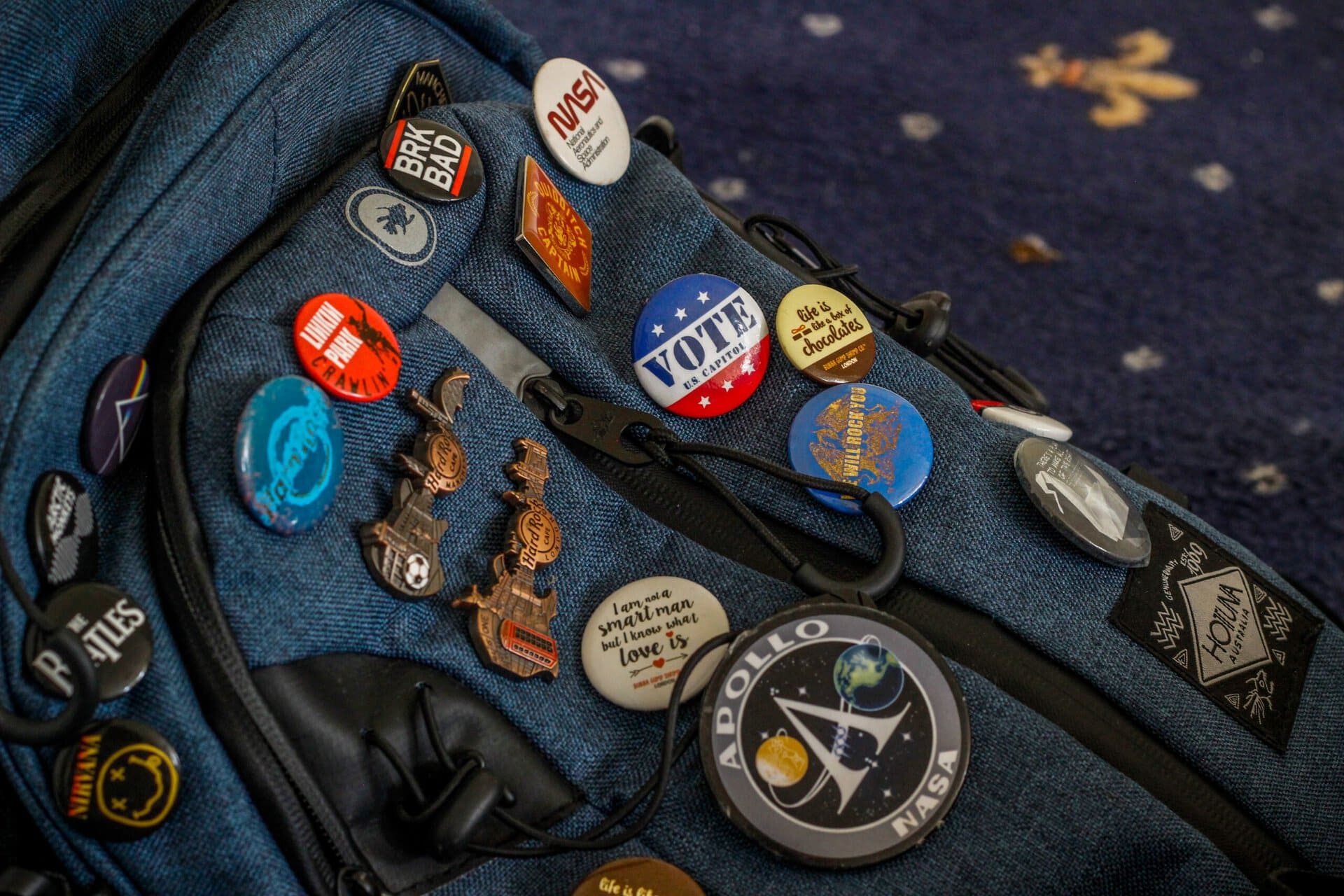 a blue backpack decorated with various colorful pins and badges, including nasa, "vote," and jurassic park designs, resting on a blue carpet with small white dots.