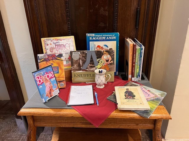 a small wooden table displays children’s books, dvds, a plush knuffle bunny, a notebook with a pen, and a red cloth in a room with wooden paneling.