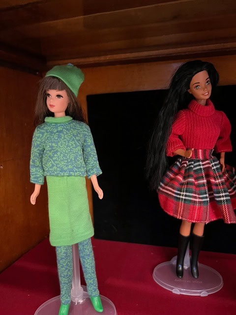 two fashion dolls on display: one has long brown hair, a green knit outfit, and hat; the other has long black hair, a red sweater, plaid skirt, and black boots.
