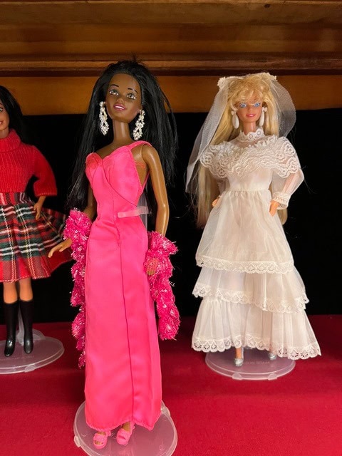 three dolls on display: the center doll wears a bright pink evening gown with earrings, the right doll wears a white bridal dress and veil, and the left doll wears a red top with a plaid skirt.