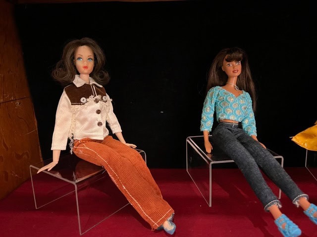 two fashion dolls are seated on clear plastic stands. one doll wears an off white and brown jacket with orange pants, and the other wears a blue patterned top with black jeans.