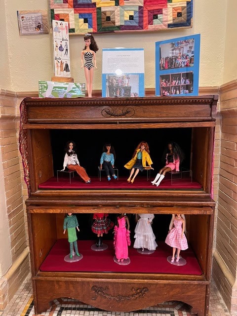 a wooden display case with shelves holds various barbie dolls in different outfits, with informational posters and a quilt displayed above it.