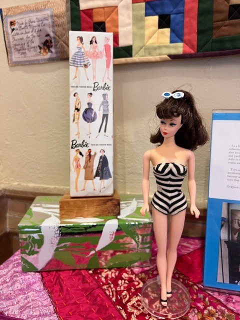 a barbie doll wearing a black and white striped swimsuit stands on display next to a stand with vintage barbie fashion illustrations.