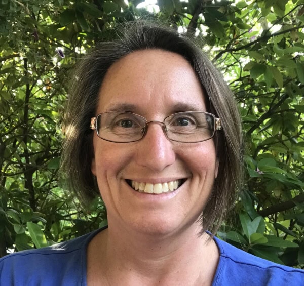 a person with short brown hair and glasses smiles outdoors in front of leafy green foliage, wearing a blue top.