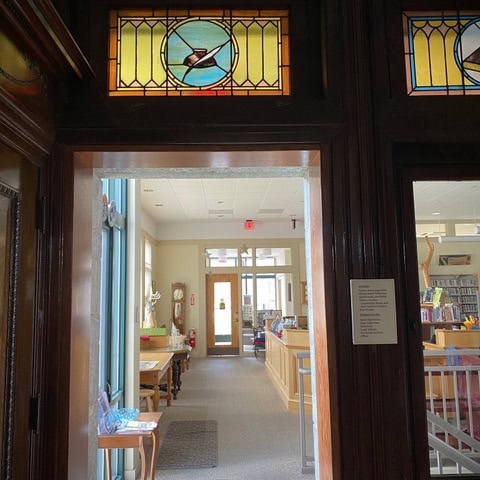 view through a doorway with stained glass artwork above, showing a brightly lit interior space with tables, bookshelves, and a fire extinguisher on the wall.