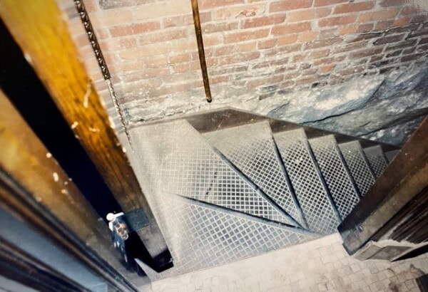 metal spiral staircase descends against a brick and stone wall, viewed from above near an open doorway, with tile flooring at the top of the stairs.