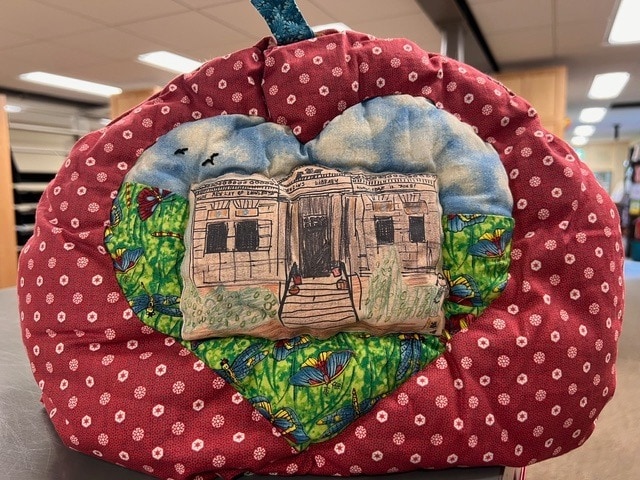 a heart shaped quilted fabric decoration with an embroidered building and landscape on the front, set against a red background with white polka dots.