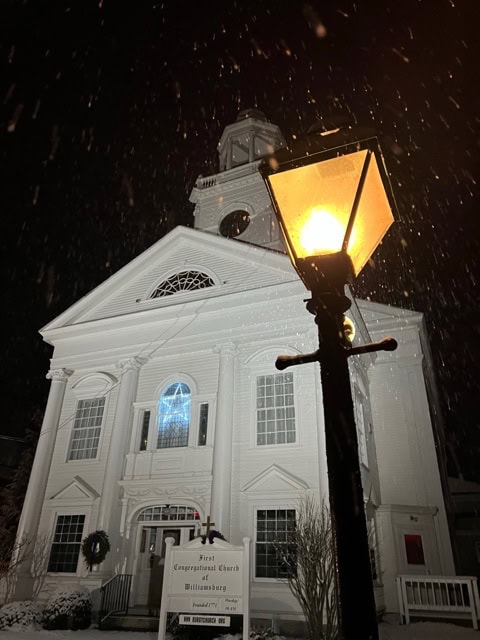 a white church building at night, illuminated by a glowing streetlamp in the foreground, with snow lightly falling.