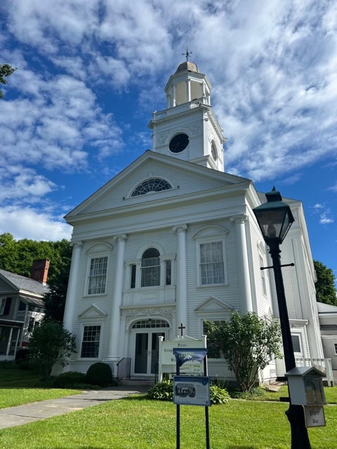 white, two story church with columns and a steeple stands on a green lawn under a partly cloudy blue sky; a sign and lamp post are visible in front.