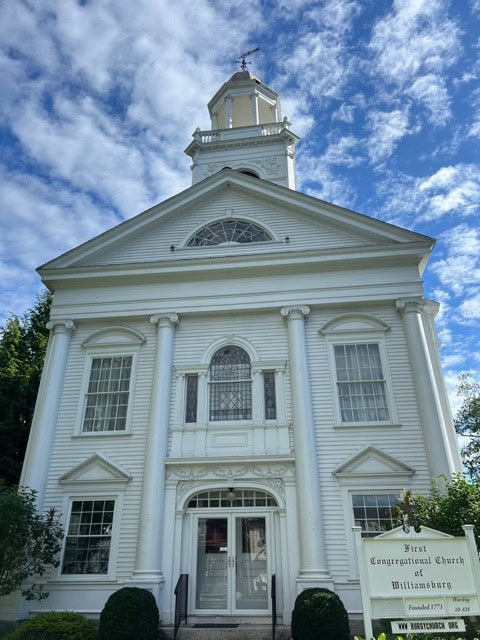 white, two story church with a tall steeple against a partly cloudy sky. a sign in front reads "first congregational church of williamsburg.