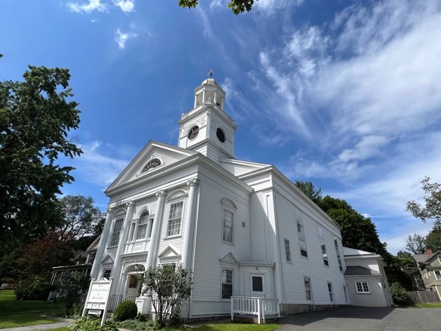 a white, two story church with a prominent steeple and columns stands under a partly cloudy blue sky, surrounded by trees and a small lawn.