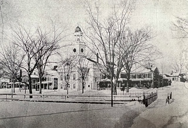 black and white photo of a snow covered town square with a central building featuring a clock tower, surrounded by bare trees and several other buildings.