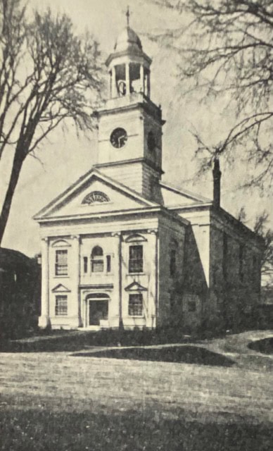 black and white photo of a historic church with a tall steeple, arched windows, and bare trees on either side.