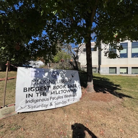 a large sign on a lawn under trees advertises the meekins library's biggest book sale during indigenous peoples weekend, with dates and times listed.
