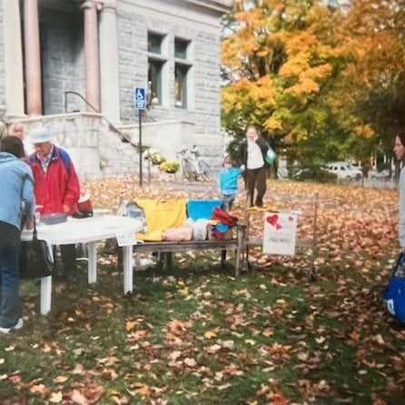 several people stand near a table with items for sale or display outside a stone building, surrounded by autumn leaves and trees.