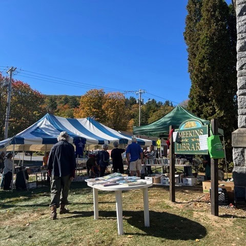 people browse tables with books at an outdoor event near the meekins library, with tents and autumn trees in the background.