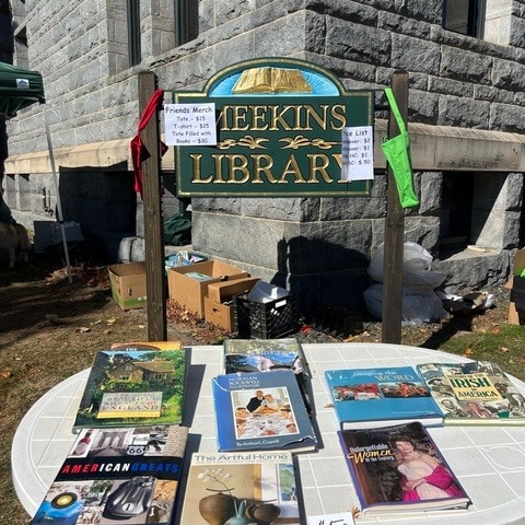 a table with books for sale is set up outside the meekins library; signs display prices for tote bags, mugs, and books under a sunny sky.