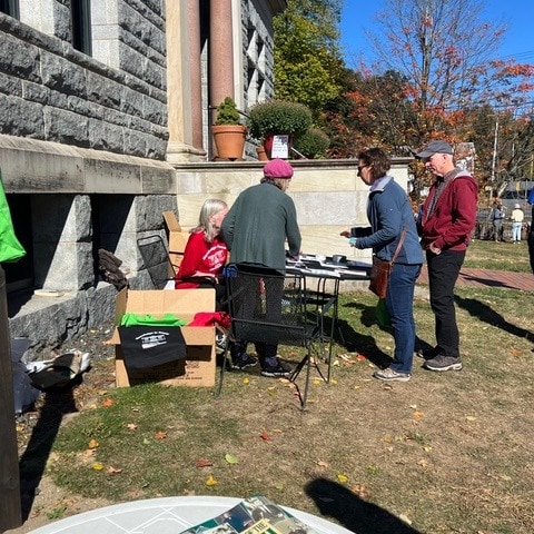 people gather at an outdoor table near a stone building, engaging with materials on the table; one person sits while others stand and interact.
