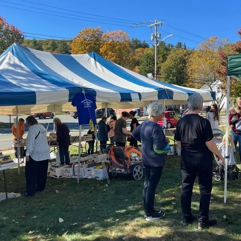people browse tables with goods under a blue and white striped tent at an outdoor market on a sunny day, with trees and cars in the background.