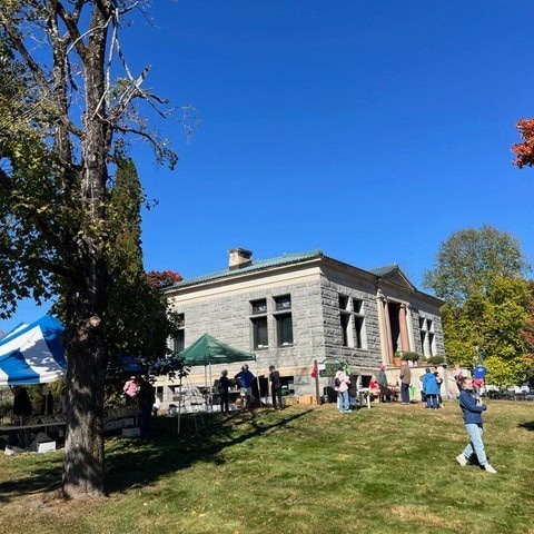 people gather outside a historic stone building with columns on a sunny day, with tents set up on the lawn and autumn trees surrounding the scene.