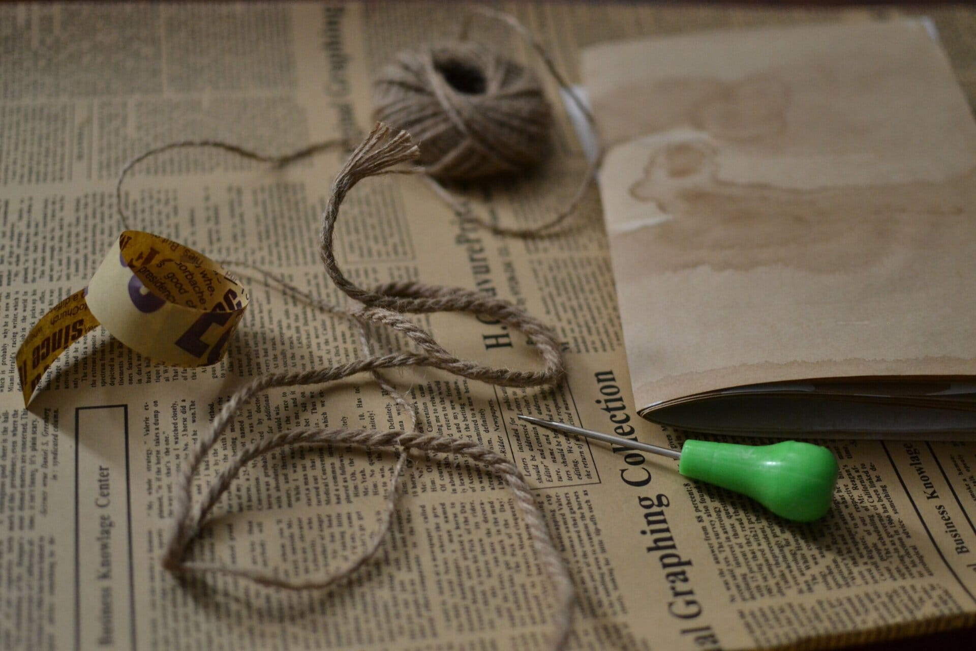 a spool of twine, a length of jute cord, and an awl with a green handle resting on a sheet of vintage newspaper.