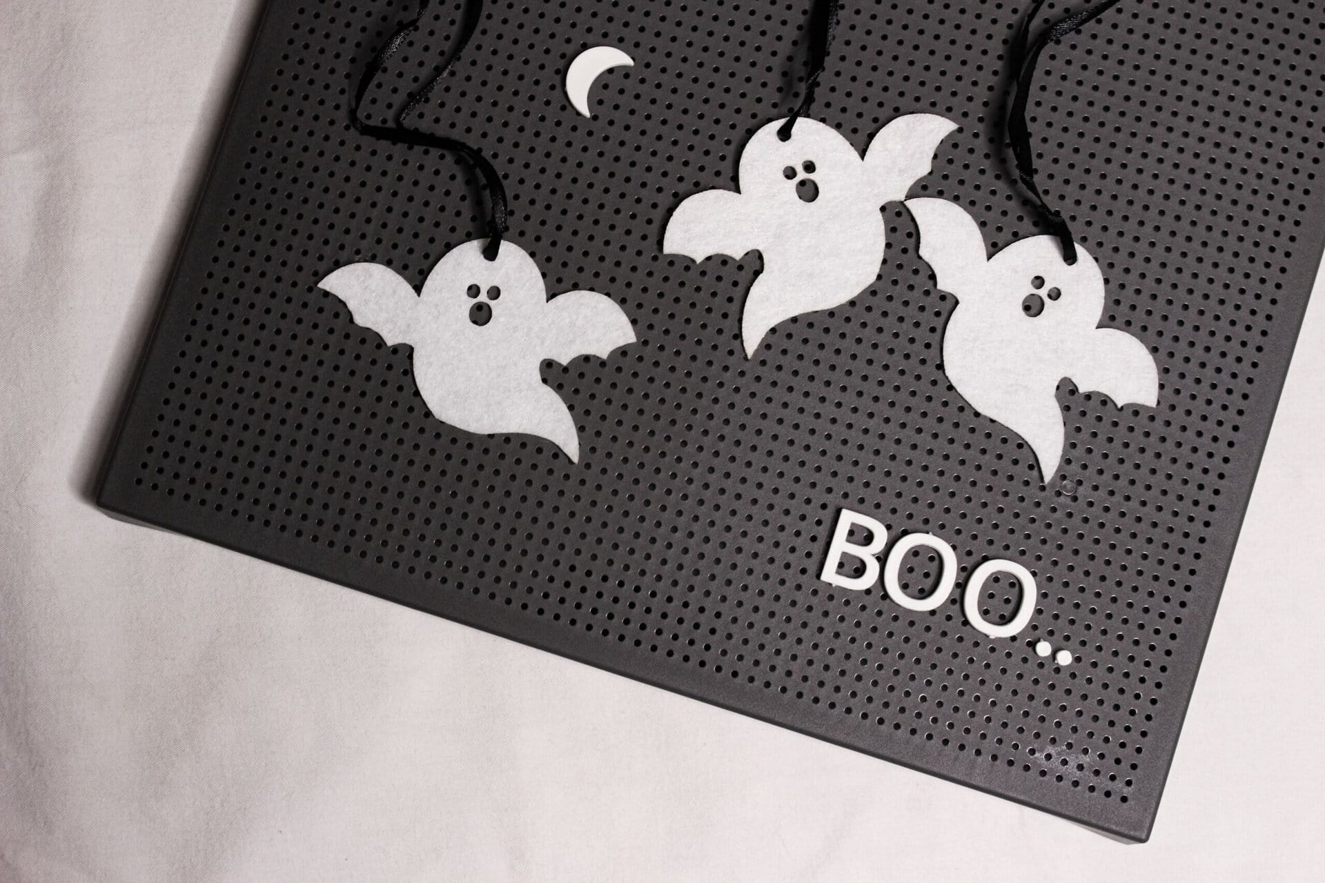 three paper ghosts and a crescent moon hang on a black pegboard, with the word “boo..” spelled out in white letters at the bottom right.