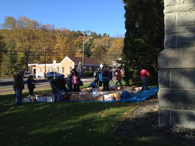 a group of people sorts through boxes on a blue tarp outdoors near a stone building, with trees and a street in the background.