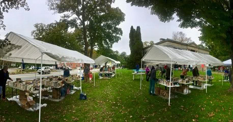 people browse tables of books under white tents at an outdoor book sale on a grassy lawn near historic buildings and trees.