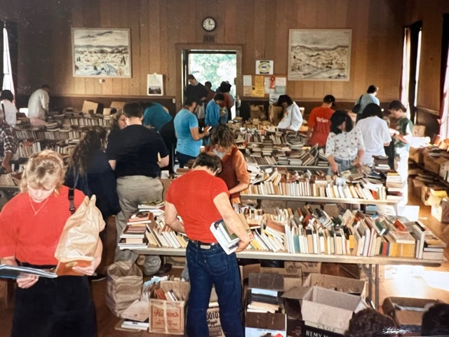 people browse through books on tables and in boxes at an indoor book sale. the room has wood paneled walls, large windows, and artwork on the walls.