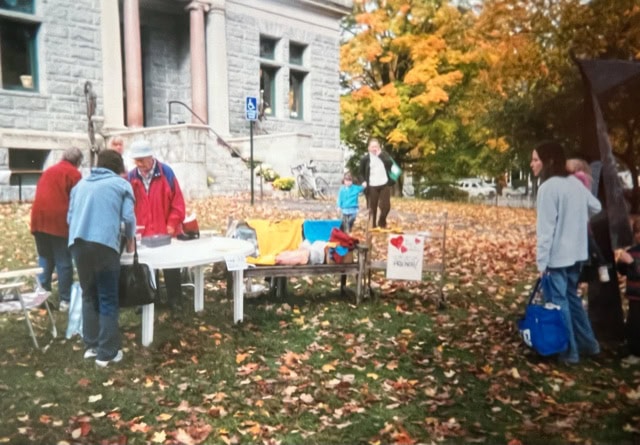 people gather around tables with various items outside a stone building on a fall day, with leaves covering the grass and trees in the background.