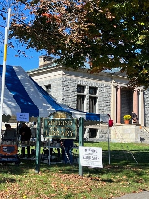 a stone library building with columns is behind a blue and white tent. signs in front read "meekins library" and "friends book sale." trees with autumn leaves are visible.