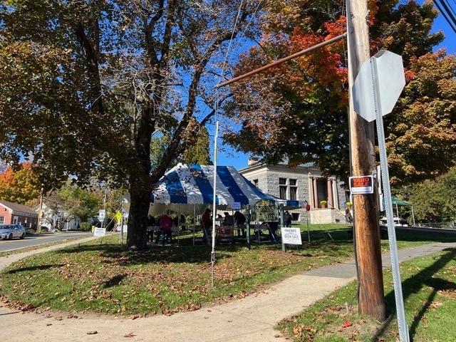 a blue and white striped tent is set up on a grassy area under trees near a stone building, with people gathered underneath and signs posted nearby.