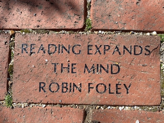 a brick engraved with the words: "reading expands the mind robin foley," set among other red bricks with some moss and pebbles.