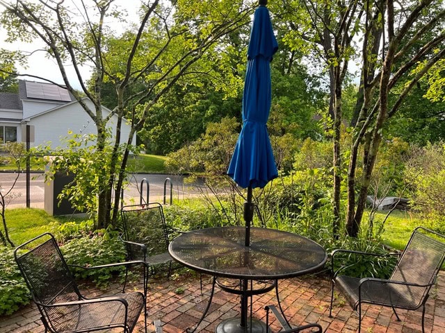 a round metal patio table with four chairs and a closed blue umbrella is set on a brick patio surrounded by greenery and trees, with a street and house in the background.