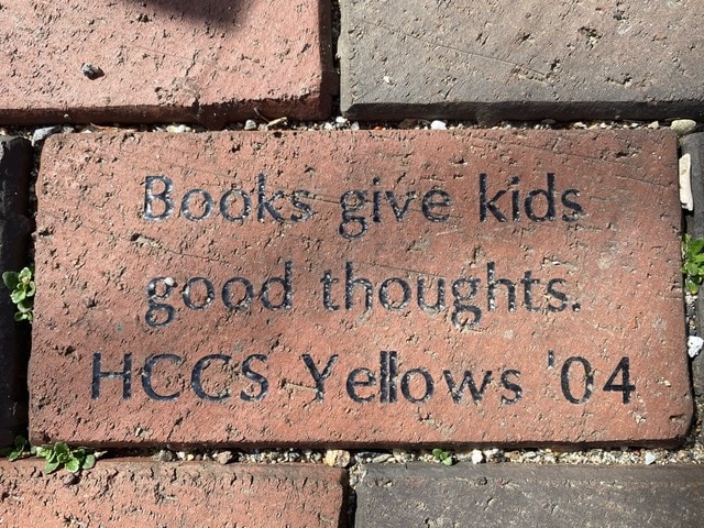 engraved brick reads: "books give kids good thoughts. hccs yellows '04," set among other bricks with small plants nearby.