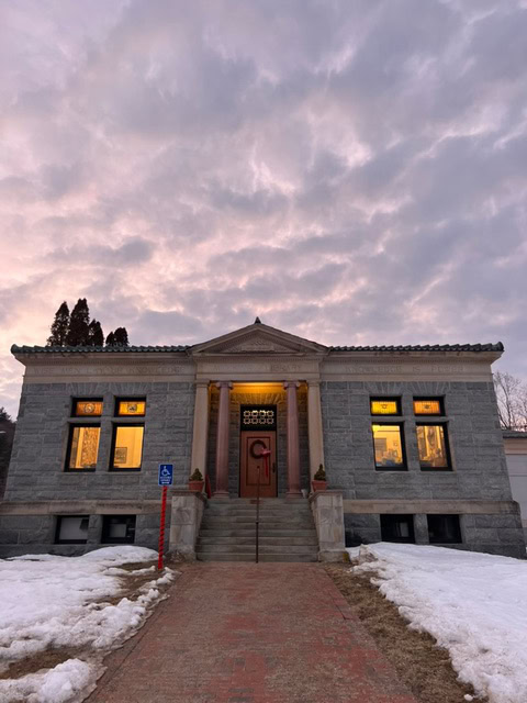 a stone building with columns and illuminated windows, set beneath a cloudy evening sky, with patches of snow and a brick walkway leading to the entrance.