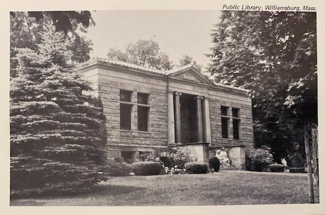 black and white photo of the public library in williamsburg, massachusetts, showing a stone building with columns surrounded by trees and landscaped bushes.