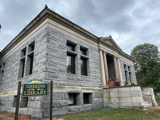 a stone building with columns and arched windows, identified by a sign in front as meekins library, stands under a cloudy sky.