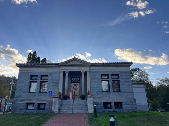 a small stone library building with columns at the entrance sits under a blue sky with clouds and sun rays. steps and a brick path lead to the front door.