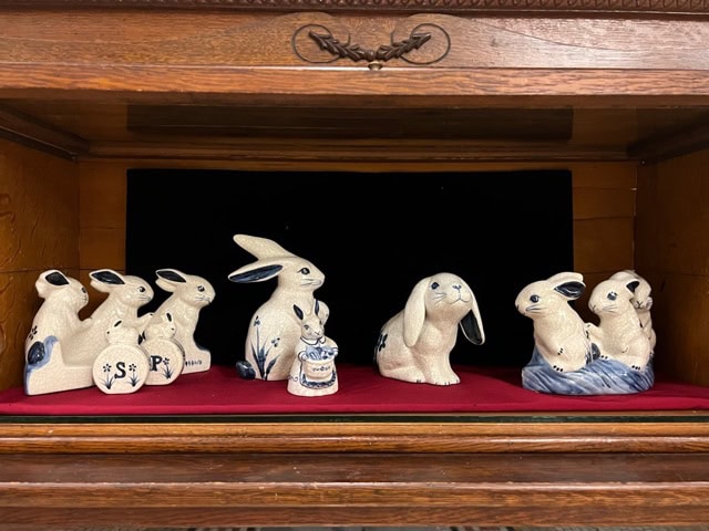 a collection of ceramic rabbit figurines, varying in size and pose, is displayed on a red cloth inside a wooden cabinet.