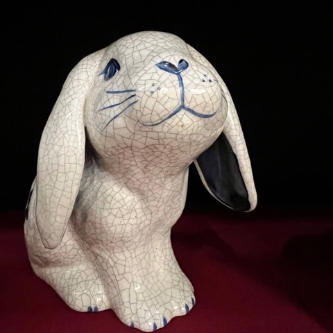 a ceramic rabbit figurine with a crackled glaze finish, blue painted details, and one ear flopped downward, set against a dark background and red surface.