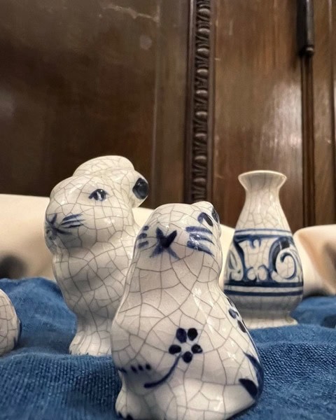 two white ceramic animal figurines and a small blue and white vase are displayed on a blue cloth, with a wooden background behind them.
