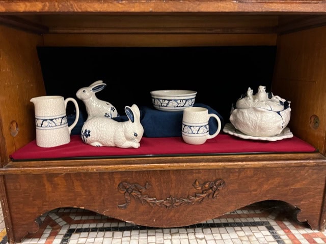 collection of ceramic dishes and figurines, including two rabbit figurines, a pitcher, mug, bowl, and a lidded dish, displayed on a red cloth inside a wooden cabinet.