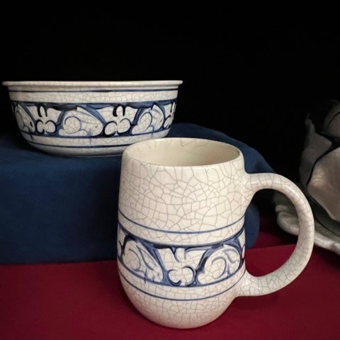 a white ceramic mug and bowl with a blue decorative pattern and crackle glaze sit on a blue and red surface.