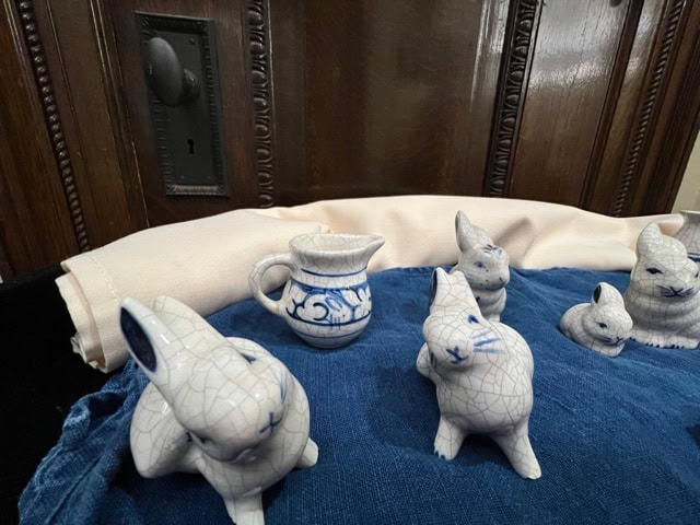 several small white ceramic rabbit figurines and a blue and white pitcher are arranged on a blue cloth in front of a wooden background.