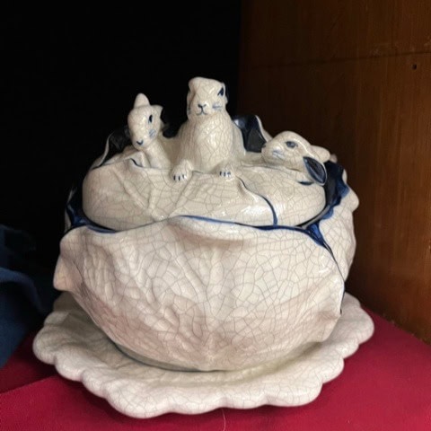 a ceramic soup tureen with a lid shaped like three rabbits sitting on cabbage leaves, displayed on a matching plate.