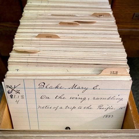 a drawer of handwritten index cards, with the front card labeled "blake, mary e. on the wing; rambling notes of a trip to the pacific. 1883." tabs separate sections alphabetically.